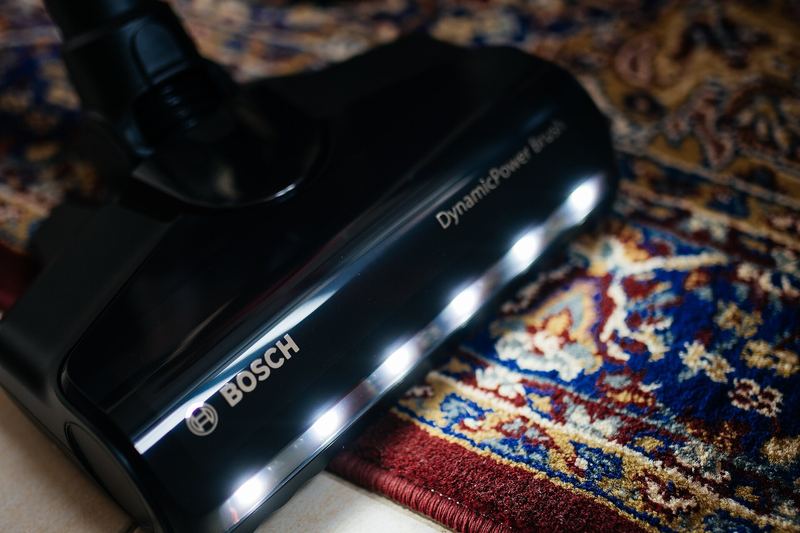 The vacuum cleaner glides over an ornate rug, illuminating its intricate patterns with bright lights. The warm ambiance of the living room enhances the cleaning